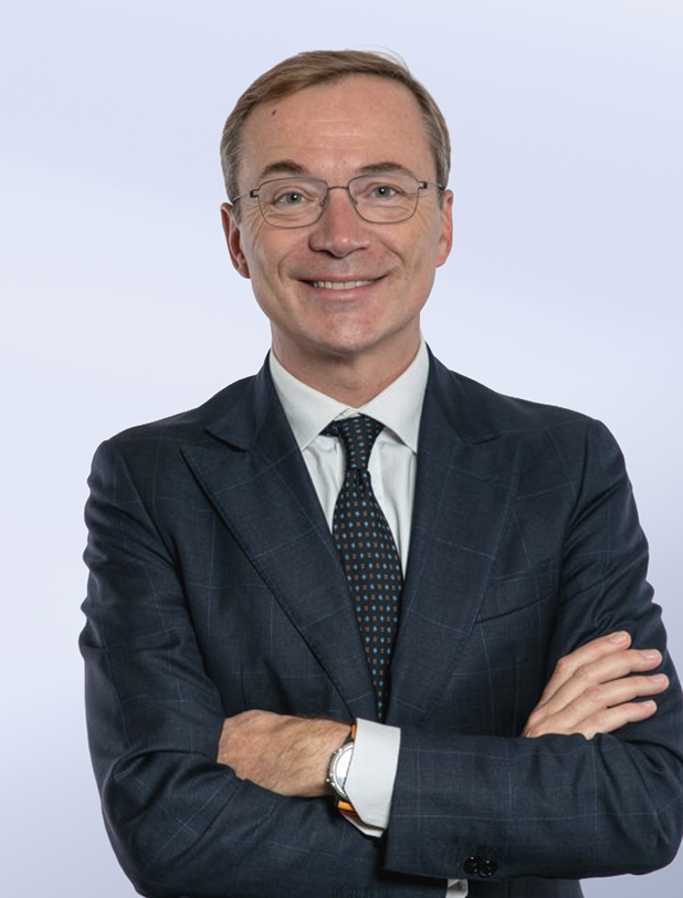 Uomo sorridente con occhiali indossa abito scuro e cravatta e posa con le braccia incrociate su sfondo chiaro.