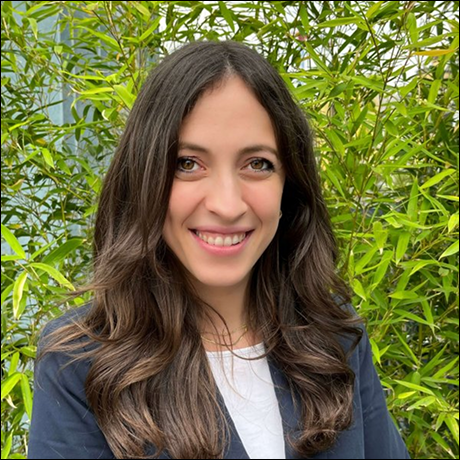 Ritratto di donna sorridente con capelli lunghi e mossi, indossa una giacca blu su maglietta bianca, con sfondo di vegetazione verde.