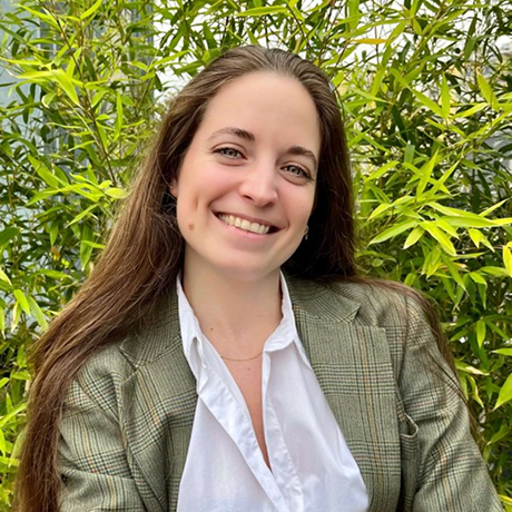 Donna sorridente con capelli lunghi e giacca a quadri verde davanti a un cespuglio verde.