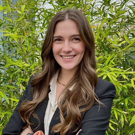 Donna sorridente con capelli lunghi e mossi, indossa giacca nera e camicia bianca, davanti a foglie verdi.