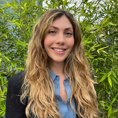 Donna bionda con capelli lunghi e ondulati, sorridente, indossa una camicia azzurra sotto una giacca nera davanti a piante verdi.