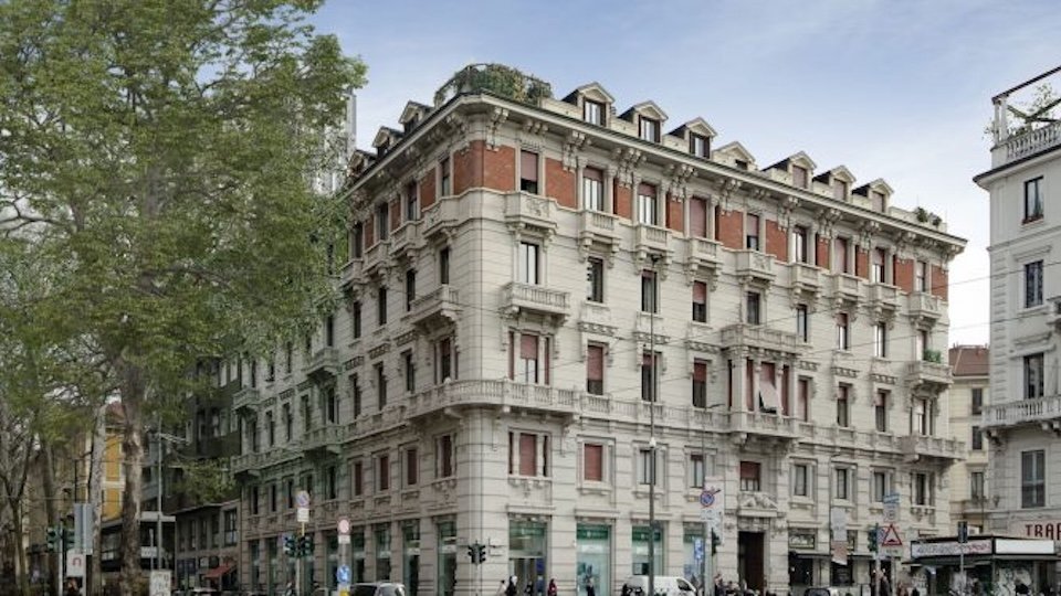 Edificio storico a Milano con facciata beige e mattoni rossi, circondato da alberi e traffico urbano.